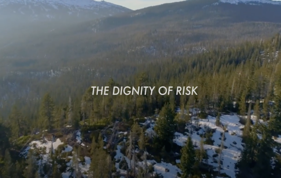 text: 'The Dignity of Risk' the background is light snow coverage on the ground with the camera positioned high in the sky offering a bird's eye view of the mountain landscape